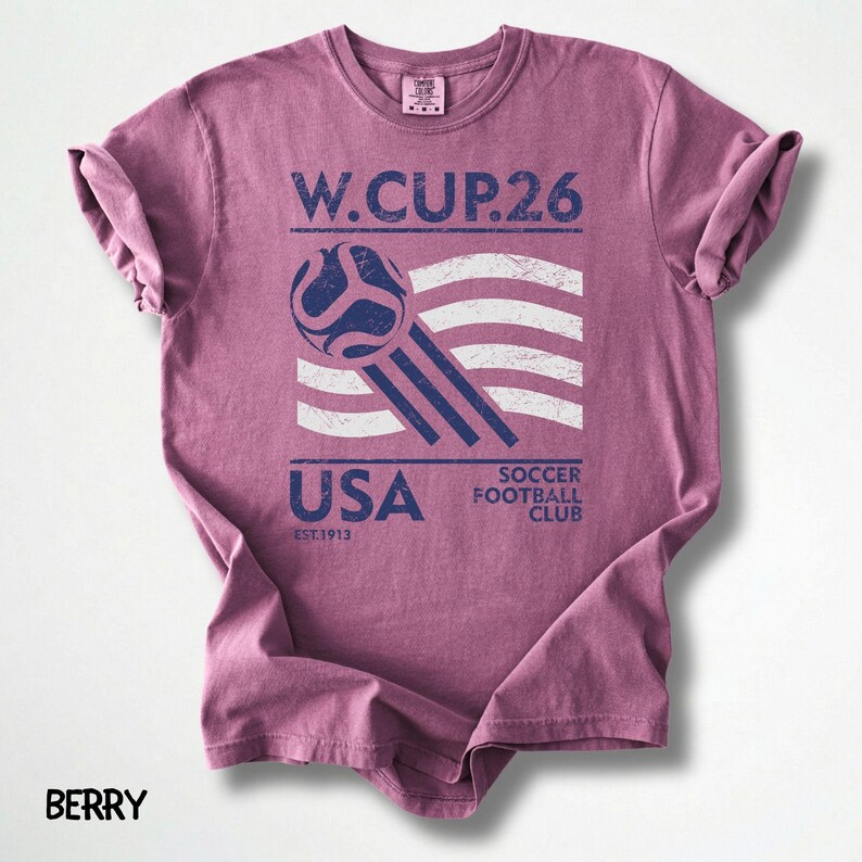 May include: Berry-colored t-shirt with a distressed graphic. The design features a soccer ball, the text "W. CUP.26", and "USA" with "Soccer Football Club" and "EST.1913". The shirt has a crew neck and rolled sleeves.
