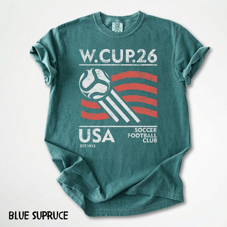 May include: Blue spruce t-shirt with a distressed graphic. The graphic includes the text "W.CUP.26", a soccer ball, and the word "USA" with "EST.1913". The shirt also says "SOCCER FOOTBALL CLUB".