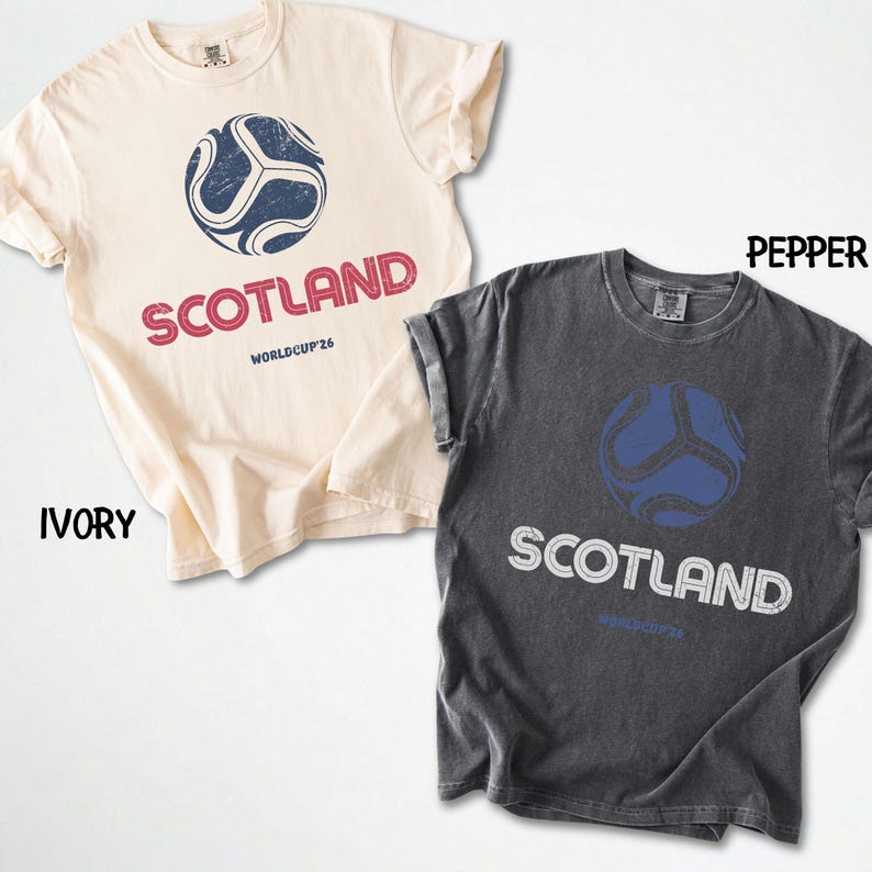 May include: Two t-shirts, one ivory and one pepper, featuring a soccer ball design. The ivory shirt displays "SCOTLAND" in red, while the pepper shirt has white text. Both shirts include "WORLDCUP'26" text below the main text.