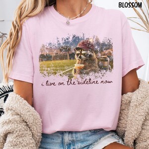 Pode incluir: Camiseta rosa claro com um guaxinim usando um bon&eacute; de beisebol e bebendo de uma x&iacute;cara, com o texto "i live on the sideline now". O guaxinim est&aacute; em um fundo de campo de futebol.