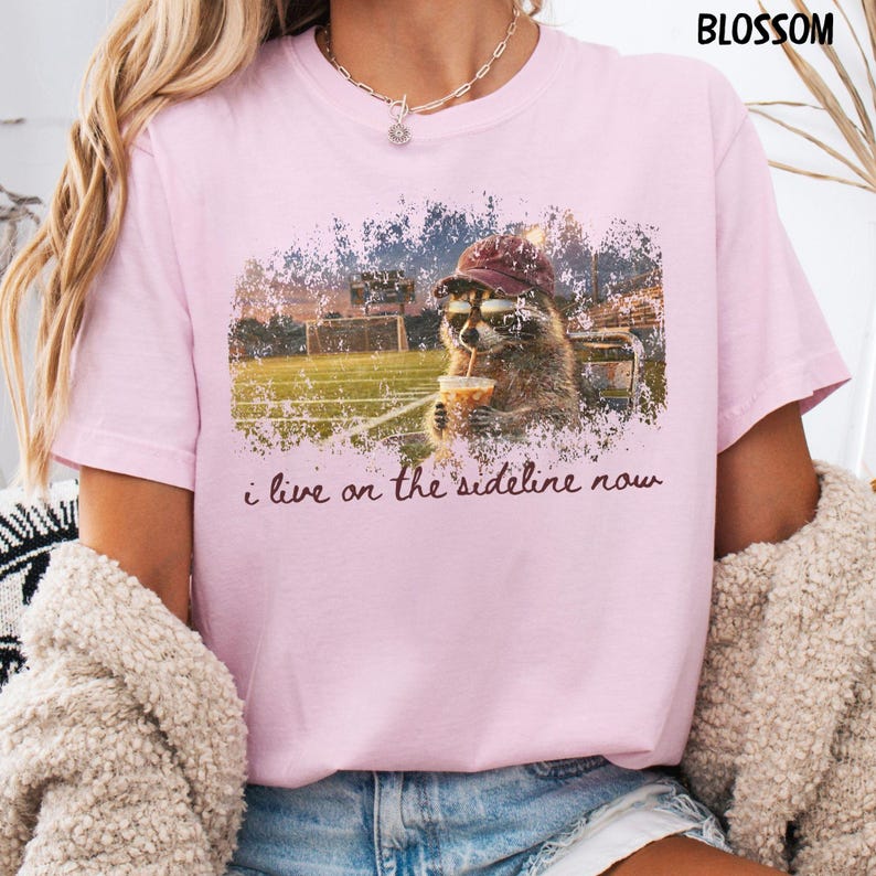 Pode incluir: Camiseta rosa claro com um guaxinim usando um bon&eacute; de beisebol e bebendo de uma x&iacute;cara, com o texto "i live on the sideline now". O guaxinim est&aacute; em um fundo de campo de futebol.