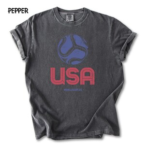 May include: Dark grey t-shirt featuring a distressed graphic of a blue and red football, the word "USA" in red, and "WORLDCUP 26" in red. The shirt has rolled sleeves and a crew neck.