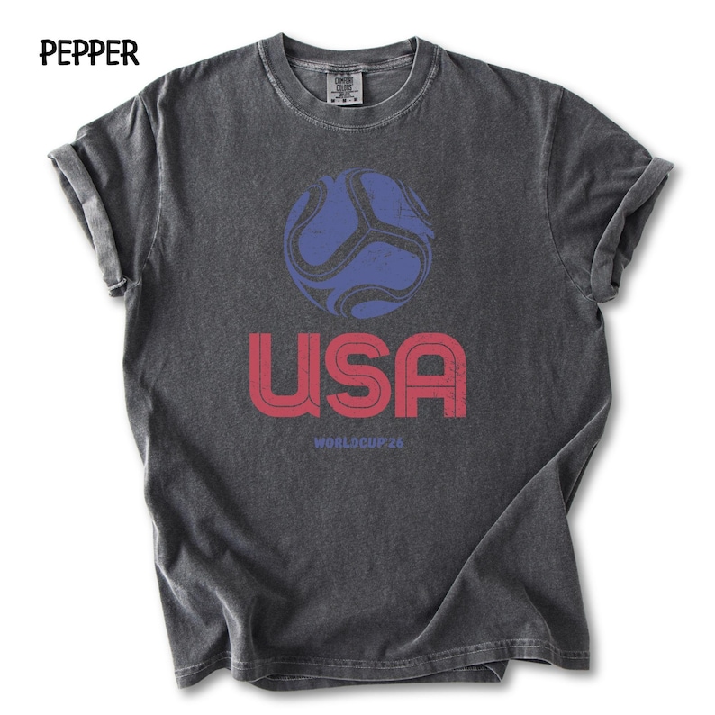 May include: Dark grey t-shirt featuring a distressed graphic of a blue and red football, the word "USA" in red, and "WORLDCUP 26" in red. The shirt has rolled sleeves and a crew neck.