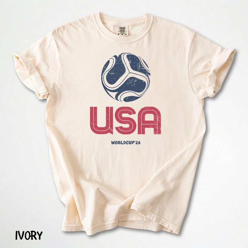 May include: Ivory t-shirt with a distressed navy and white football graphic above the red text "USA" and "WORLDCUP'26". The shirt is made of soft material and has rolled sleeves.