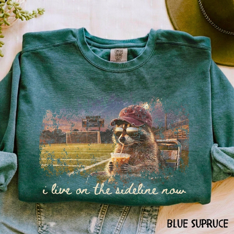 Pode incluir: Camisola azul abeto com um guaxinim a usar &oacute;culos de sol e bon&eacute;, a beber de um copo. O design inclui o texto "i live on the sideline now" e um campo de futebol. A camisola est&aacute; rotulada como "BLUE SPRUCE".