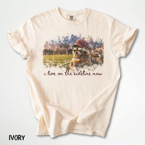 Pode incluir: Camiseta cor marfim com um gr&aacute;fico de um guaxinim usando &oacute;culos de sol e um chap&eacute;u, bebendo de um canudo. O gr&aacute;fico &eacute; colocado em um fundo desfocado de um campo esportivo, com o texto "i live on the sideline now".