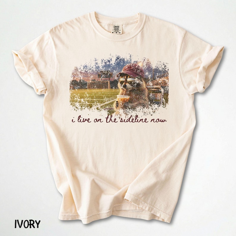 Pode incluir: Camiseta cor marfim com um gr&aacute;fico de um guaxinim usando &oacute;culos de sol e um chap&eacute;u, bebendo de um canudo. O gr&aacute;fico &eacute; colocado em um fundo desfocado de um campo esportivo, com o texto "i live on the sideline now".