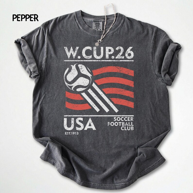 Flat lay of a pepper dark gray Comfort Colors shirt with a white and red distressed USA Soccer 2026 print and soccer ball icon.