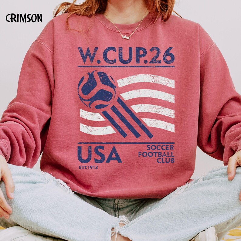 May include: Crimson-colored sweatshirt with a graphic featuring a soccer ball, the American flag, and the text "W. CUP. 26 USA SOCCER FOOTBALL CLUB EST. 1913". The sweatshirt is a casual style.