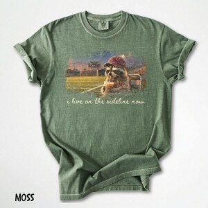 Pode incluir: Camiseta verde musgo com um guaxinim usando um chap&eacute;u e bebendo de um copo, com o texto "i live on the sideline now". O guaxinim est&aacute; em um est&aacute;dio. A camisa &eacute; feita de um material macio.