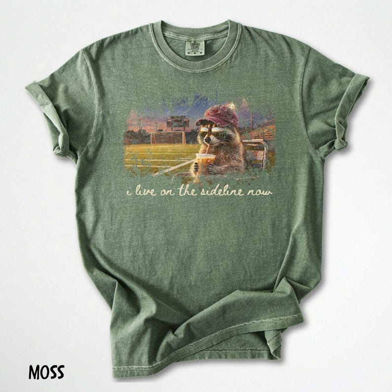 Pode incluir: Camiseta verde musgo com um guaxinim usando um chap&eacute;u e bebendo de um copo, com o texto "i live on the sideline now". O guaxinim est&aacute; em um est&aacute;dio. A camisa &eacute; feita de um material macio.