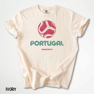 May include: Cream-coloured t-shirt with a distressed red and white football graphic. Below the graphic, the word "PORTUGAL" is printed in teal, with "WORLDCUP'26" underneath. The word "IVORY" is printed at the bottom.