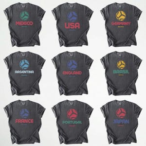 May include: Nine dark grey t-shirts, each displaying a country name and a soccer ball design. The shirts feature the names Mexico, USA, Germany, Argentina, England, Brazil, France, Portugal, and Japan, each with a unique ball colour.
