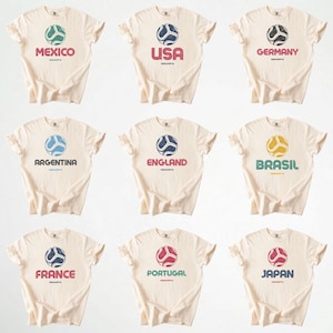 May include: Nine cream-coloured t-shirts, each displaying a country name and a soccer ball design. The shirts feature the names "Mexico", "USA", "Germany", "Argentina", "England", "Brasil", "France", "Portugal", and "Japan" in various colours.