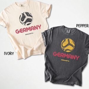 May include: Two t-shirts, one ivory and one pepper, with a soccer ball design. The word "GERMANY" is printed in red below the ball, with "WORLDCUP'26" underneath. The shirts are short-sleeved and have a vintage, worn appearance.