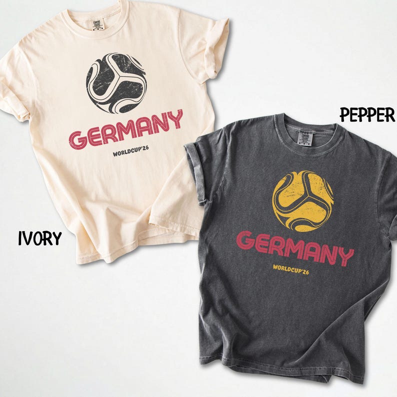 May include: Two t-shirts, one ivory and one pepper, with a soccer ball design. The word "GERMANY" is printed in red below the ball, with "WORLDCUP'26" underneath. The shirts are short-sleeved and have a vintage, worn appearance.