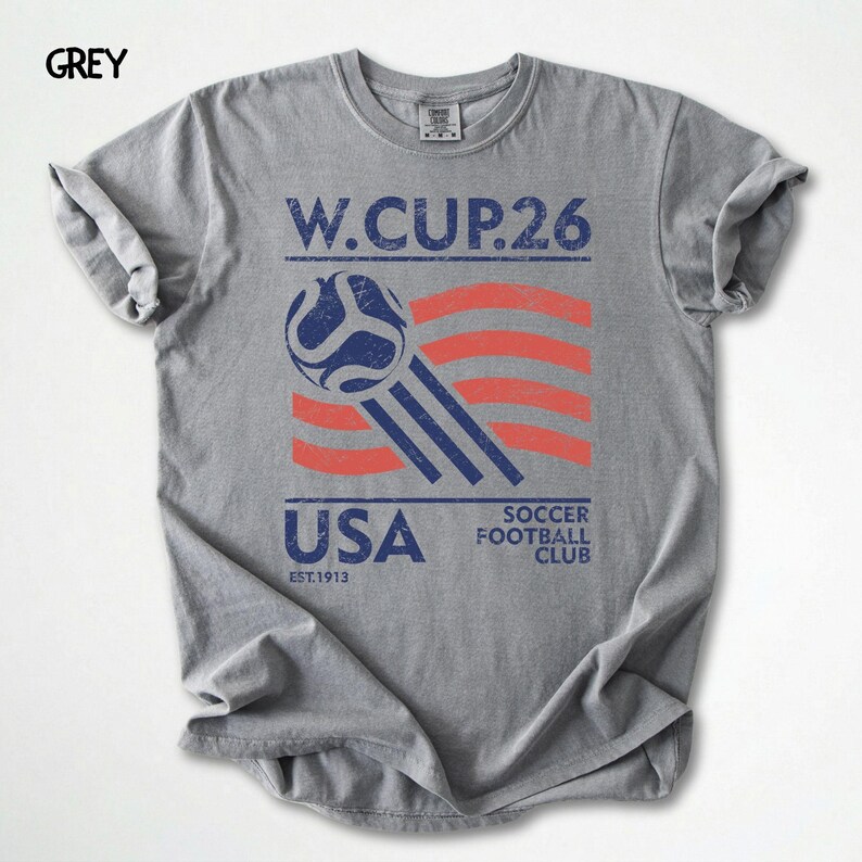 May include: A grey t-shirt with a distressed graphic featuring a soccer ball, red and white stripes, and the text "W.CUP.26 USA SOCCER FOOTBALL CLUB EST.1913". The shirt has rolled-up sleeves.