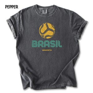 May include: Charcoal grey t-shirt with a distressed print. It features a yellow and black football design above the word "BRASIL" in teal, with "WORLDCUP '26" below. The shirt has rolled-up sleeves and the word "PEPPER" at the top.