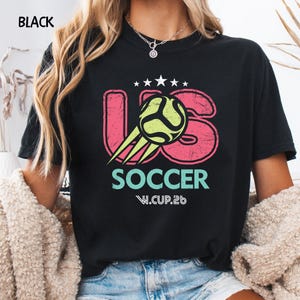 Model wearing a black Comfort Colors 1717 t-shirt with a distressed US Soccer W.CUP.26 graphic print on the front.