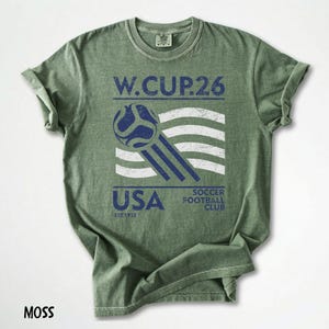 May include: Moss green t-shirt with a distressed graphic. The graphic includes the text "W.CUP.26", a soccer ball, and the word "USA" with "SOCCER FOOTBALL CLUB" below. The shirt has rolled-up sleeves.