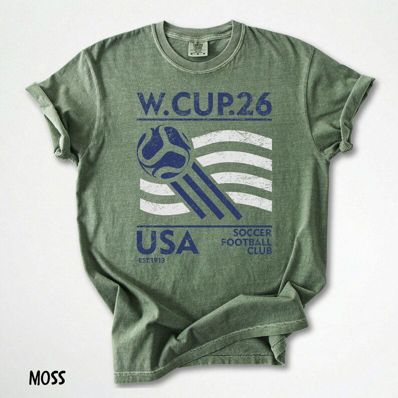 May include: Moss green t-shirt with a distressed graphic. The graphic includes the text "W.CUP.26", a soccer ball, and the word "USA" with "SOCCER FOOTBALL CLUB" below. The shirt has rolled-up sleeves.