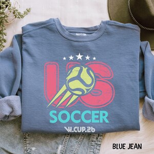 A folded blue jean color sweatshirt with a vintage style US Soccer ball and stars design in pink and lime green.