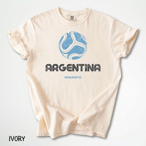 May include: Ivory t-shirt with a distressed blue soccer ball graphic and the word "ARGENTINA" in a textured font. Below the text is "WORLDCUP'26". The word "IVORY" is printed at the bottom of the shirt.