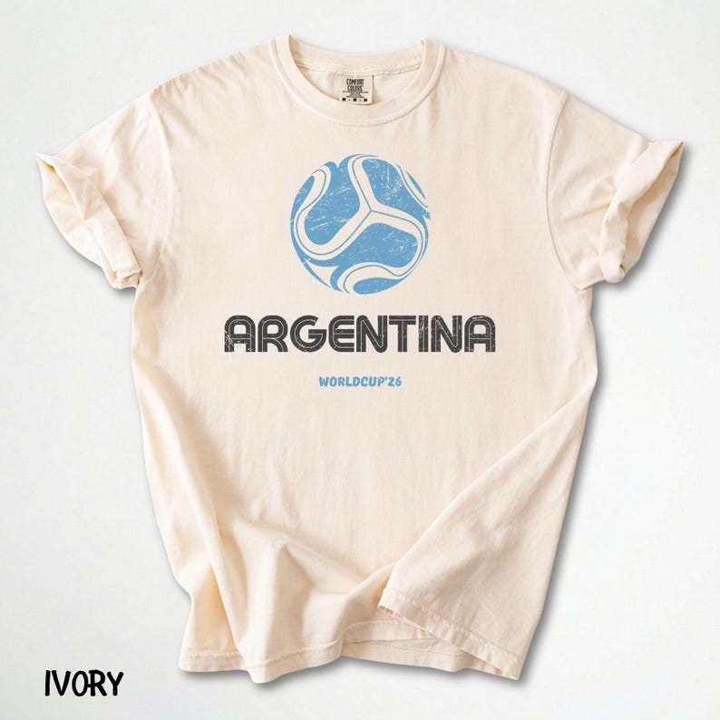 May include: Ivory t-shirt with a distressed blue soccer ball graphic and the word "ARGENTINA" in a textured font. Below the text is "WORLDCUP'26". The word "IVORY" is printed at the bottom of the shirt.