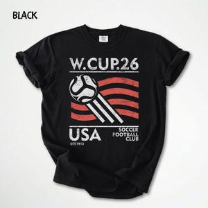 May include: Black t-shirt with a distressed graphic featuring a soccer ball, red and white stripes, and the text "W.CUP.26 USA SOCCER FOOTBALL CLUB EST.1913". The word "BLACK" is printed at the top left.