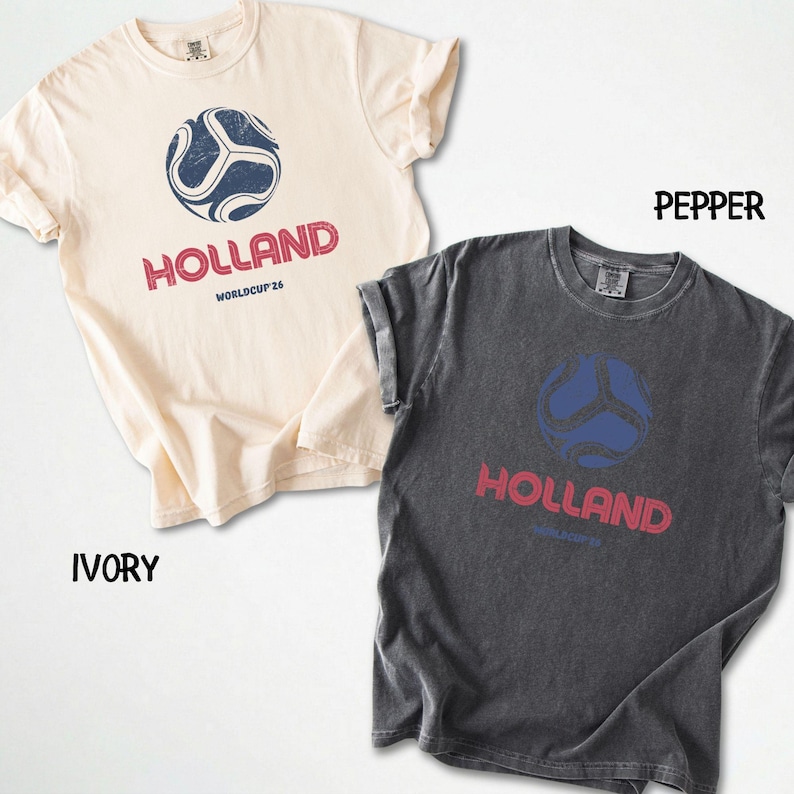 May include: Two t-shirts, one ivory and one pepper, with a distressed print. Each shirt features a blue soccer ball graphic above the word "HOLLAND" in red, with "WORLDCUP 26" below. The shirts have rolled sleeves.