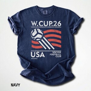 Flat lay of a navy blue t-shirt featuring a high-contrast white and red retro soccer 26 graphic with a weathered American flag aesthetic.