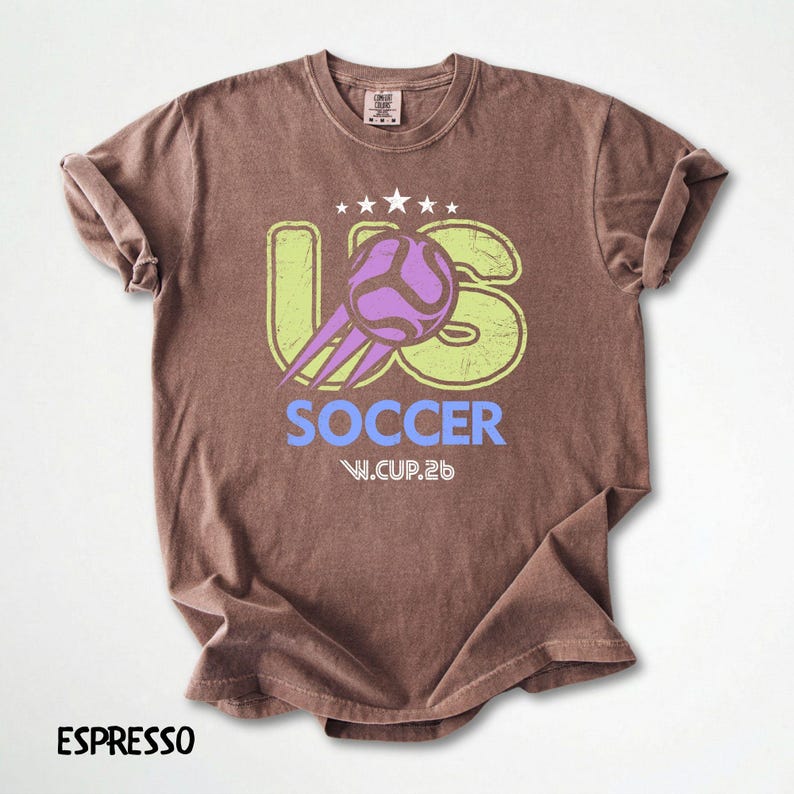 Flat lay of an espresso brown t-shirt showing a distressed US Soccer 26 print with purple and lime accents.