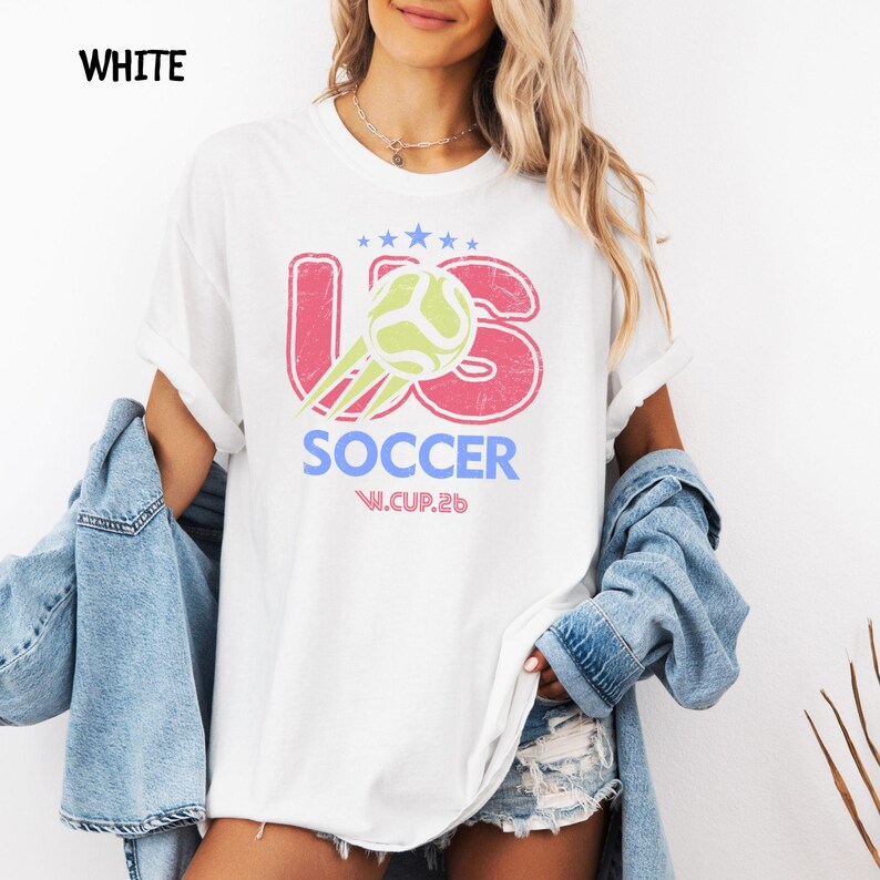 Model wearing a crisp white t-shirt with a vibrant US Soccer 2026 graphic print, styled with a denim jacket.