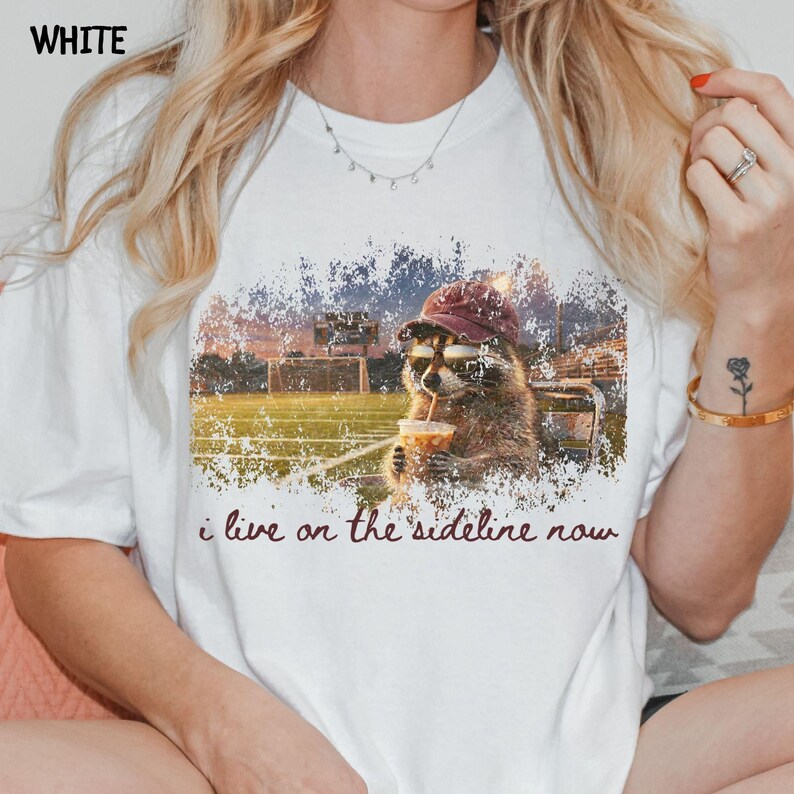 Pode incluir: Camiseta branca com um guaxinim usando &oacute;culos de sol e um chap&eacute;u bord&ocirc;, segurando uma bebida. O design inclui um campo de futebol e o texto "i live on the sideline now."