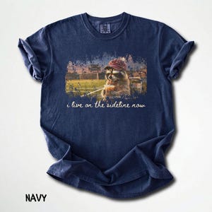 Pode incluir: Camiseta azul marinho com um desenho de um guaxinim usando &oacute;culos de sol e chap&eacute;u, segurando uma bebida com canudo, com o texto "i live on the sideline now". A imagem est&aacute; em um campo esportivo.