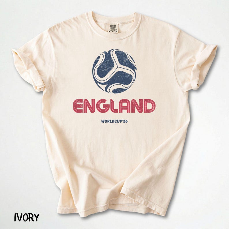 May include: Cream-coloured t-shirt with a distressed navy and white football graphic and the word "ENGLAND" in red. Below is "WORLDCUP '26". The shirt has rolled sleeves and is made of a soft material. The word "IVORY" is at the bottom.