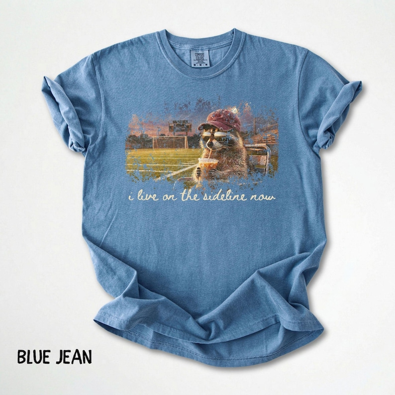 Pode incluir: Camiseta azul jeans com um guaxinim usando um bon&eacute; de beisebol e bebendo de um copo com canudo. O guaxinim est&aacute; sentado em um banco com um campo de futebol ao fundo. O texto "i live on the sideline now" est&aacute; impresso na camiseta.