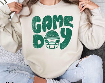 May include: A sand-colored sweatshirt with the words "GAME DAY" in distressed green lettering, featuring a football helmet graphic. The sweatshirt is a casual, comfortable style.