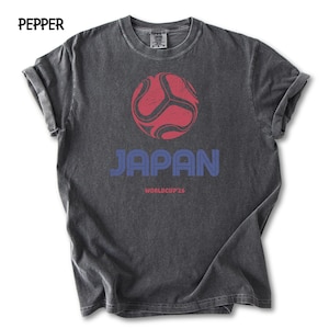 May include: A dark grey t-shirt featuring a red and black football graphic above the word "JAPAN" in blue. Below the word "JAPAN" is the text "WORLDCUP 26". The shirt has rolled-up sleeves.