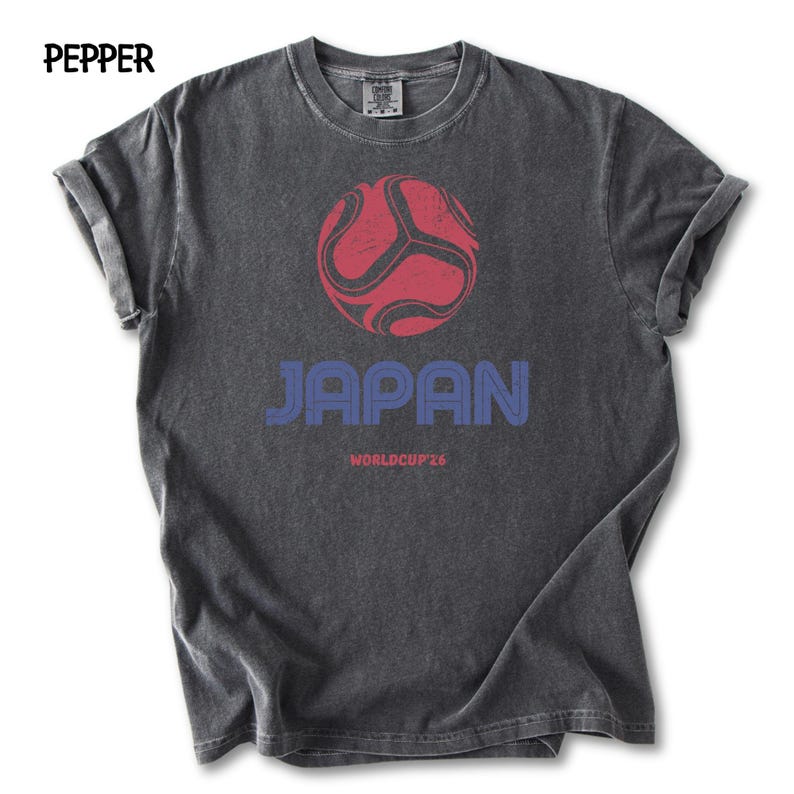 May include: A dark grey t-shirt featuring a red and black football graphic above the word "JAPAN" in blue. Below the word "JAPAN" is the text "WORLDCUP 26". The shirt has rolled-up sleeves.