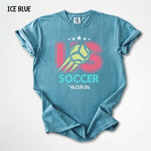 Flat lay view of an ice blue Comfort Colors 1717 shirt with a colorful vintage football world cup design.