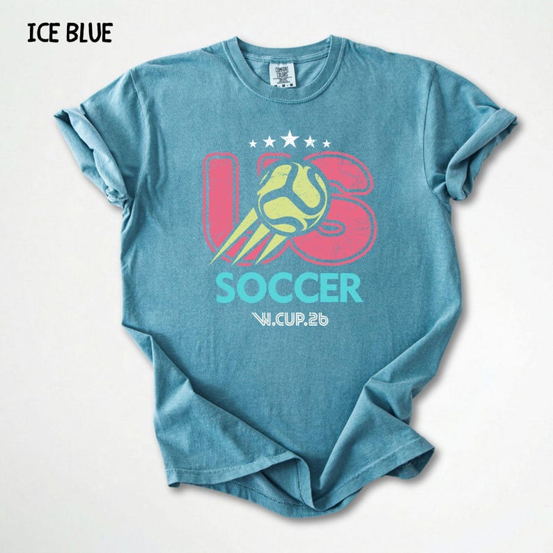 Flat lay view of an ice blue Comfort Colors 1717 shirt with a colorful vintage football world cup design.