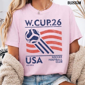 A blonde model wearing a blossom pink Gildan t-shirt with a distressed blue and red USA Soccer W.CUP.26 graphic featuring a soccer ball and waving stripes.