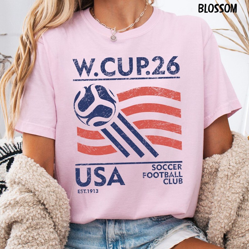 A blonde model wearing a blossom pink Gildan t-shirt with a distressed blue and red USA Soccer W.CUP.26 graphic featuring a soccer ball and waving stripes.