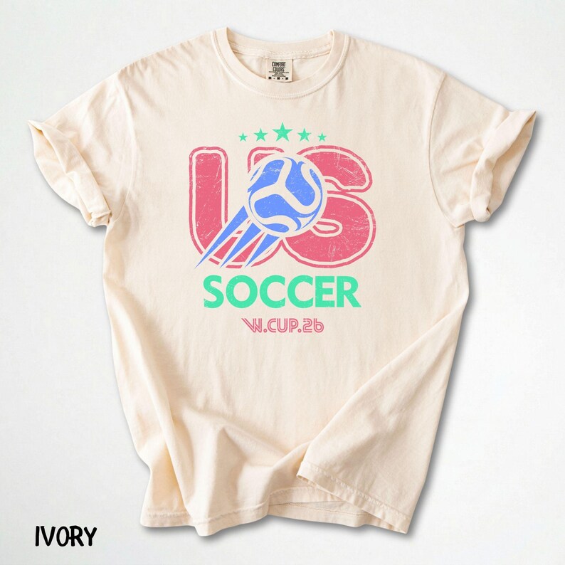 Flat lay of an ivory off-white t-shirt with a pastel-toned US Soccer 26 logo and a soft distressed texture.
