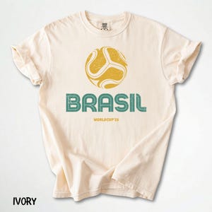 May include: Ivory t-shirt with a distressed yellow and green football graphic and the word "BRASIL" in green. Below is "WORLDCUP'26". The shirt has rolled-up sleeves and the word "IVORY" at the bottom.