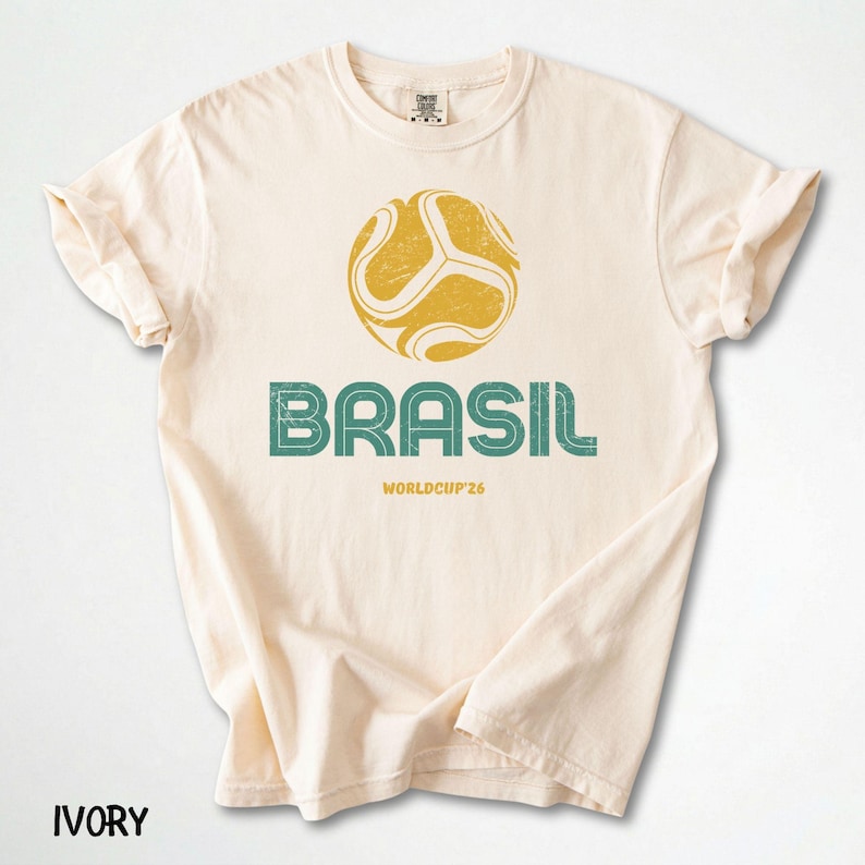 May include: Ivory t-shirt with a distressed yellow and green football graphic and the word "BRASIL" in green. Below is "WORLDCUP'26". The shirt has rolled-up sleeves and the word "IVORY" at the bottom.