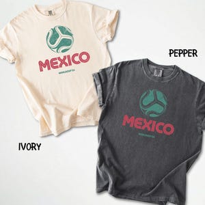 May include: Two t-shirts, one ivory and one pepper, with rolled sleeves. Each shirt features a soccer ball graphic above the word "MEXICO" in red, with "WORLDCUP26" below. The soccer ball is teal.