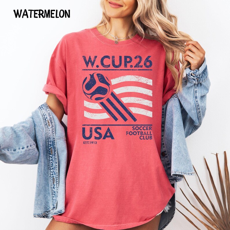 A model wearing a watermelon pink oversized t-shirt with a blue and white USA Soccer Football Club 26 graphic, styled with a denim jacket.