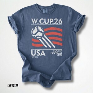 A denim blue t-shirt laid flat featuring a distressed white and red American flag themed soccer graphic for the 2026 World Cup.